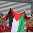 Protesters wearing read hold a Palestinian flag and cardboard signs with slogans opposing the activities of the Israeli weapons maker Elbit Systems 