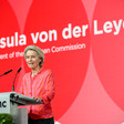 European Commission President Urusla von der Leyen stands at a podium with her name written behind her in white on a red backdrop