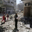 Palestinian children and adults, including a woman holding an infant, step across a rubble-strewn street