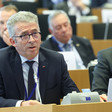 Man wearing dark suit and tie sits at desk with a microphone on one side of him and a sign saying C. Gomart on the other