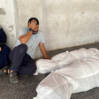 A man and a woman mourn next to two bodies wrapped in white shrouds