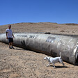 A man and a pet dog stand next to a massive missile fragment on a barren landscape