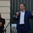 A man wearing a dark blue suit and a white shirt holds a microphone as another man stands behind him 