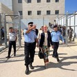 A woman stands between two men in uniform with a large building and a large gate partly topped with barbed wire behind them