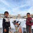 Two boys stand facing the camera in front of white tents, with destroyed buildings in the background