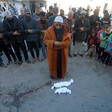A man wearing a dark orange coat prays over two tiny bodies in white shrouds. A crowd of mourners is behind him.