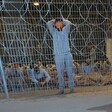 Men blindfolded and bound sit or stand behind razor wire fence