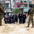 People march on a dirt road as they're watched over by two Israeli soldiers