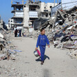 A girl in blue clothes walks past piles of rubble, carrying a pot