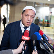 An imam wearing a white pray hat and a navy suit speaks to reporters holding microphones