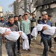 A group of men, two of them carrying small bodies wrapped in white shrouds, walk down a street