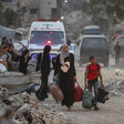 Women and children carry belongings amidst rubble. Ambulances are behind them. 
