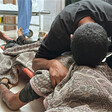 A man cradles the body of a loved one on the floor of a hospital. The body is wrapped in a blanket.