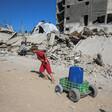 A child, dressed in red, pulls a can of water on a small cart up a hill, surrounded by rubble of destroyed buildings.