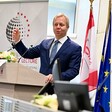 A man wearing a suit stands at a podium with flags behind him 
