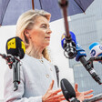 European Commission President Ursula von der Leyen stands beneath an umbrella and surrounded by television microphones 