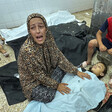 A woman wearing prayer clothes mourns over the body of a small child under a blue medical blanket. Two small children lie motionless behind her as an older boy sits next to her.