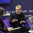 A man wearing dark clothes does a hand gesture as he sits beside another person on a stage 