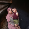 A woman wearing pink prayer clothes holds a toddler, dressed in a red shirt and green shorts. The toddler is missing his left arm.