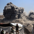 An enormous plume of black smoke rises against a blue sky, near people watching from their tent shelters.