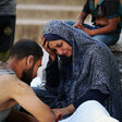 A woman in a blue headscarf holds her head in her hand. A man faces her. They are both looking at a body wrapped in a white shroud.