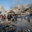 People stand on top of the rubble of a destroyed building, with black smoke rising around them. Several small fires are visible in front of them.