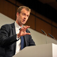 A man wearing a navy suit and white shirt stands at a podium