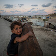A small child clings to a rusty metal object, looking at the camera. Tent shelters are in the foreground.