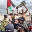 A group of young Palestinian men and children stand in a group. Carried on shoulders, two men hold a Palestinian flag and a symbolic key. Everyone is smiling. 