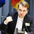 A man wearing a dark jacket and a white shirt sits at a desk with a microphone on it