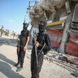 Two armed men stand guard in front of damaged buildings