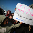 A group of women and youth stand together and hold a sign that reads "No more tents in winter hell, let caravans in now!"