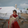 A girl stands in front of a tent shelter. She is dressed in red and wrapped in a blanket