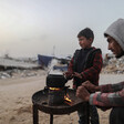 A man and a boy warm their hands over a small tray with a fire and a teapot over it