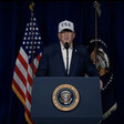 Donald Trump stands at presidential podium speaking and wearing a baseball hat reading "USA"