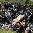 A large crowd of people gather around a body, carried on a stretcher, wrapped in a brown blanket.