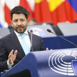 Man wearing a dark suit and white shirt stands behind a podium bearing the European Parliament logo 
