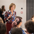 Katharina von Shnurbein, wearing floral print blouse, holds microphone and stands amid a group of sitting people in conference room