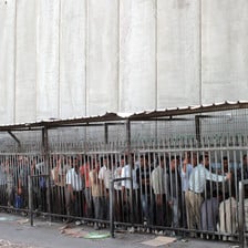 Palestinian men queue at checkpoint next to very tall concrete wall