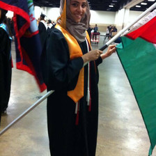 Young woman wearing graduation gown smiles while holding Palestinian flag