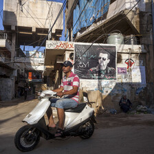 Scene shows youth on scooter riding past wall covered with poster and graffiti