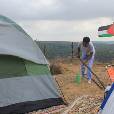 Man picks up bar amid tents with Palestine flag and landscape in background