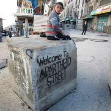 Boy sits on cement block which is spray painted with a stencil reading Welcome to apartheid street