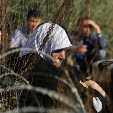 Elderly woman sits next to suitcase near fence and barbed wire