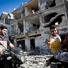 Smiling young men on motorbikes give victory sign in front of shelled building in Gaza City