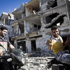 Smiling young men on motorbikes give victory sign in front of shelled building in Gaza City