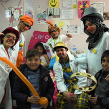 Men and women in silly hats and wigs wearing red foam noses pose with children in hospital room