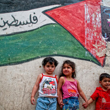 Small children stand against wall with mural of Palestine flag