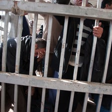 Crowd of men stand or sit behind metal bars at checkpoint