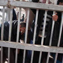 Crowd of men stand or sit behind metal bars at checkpoint